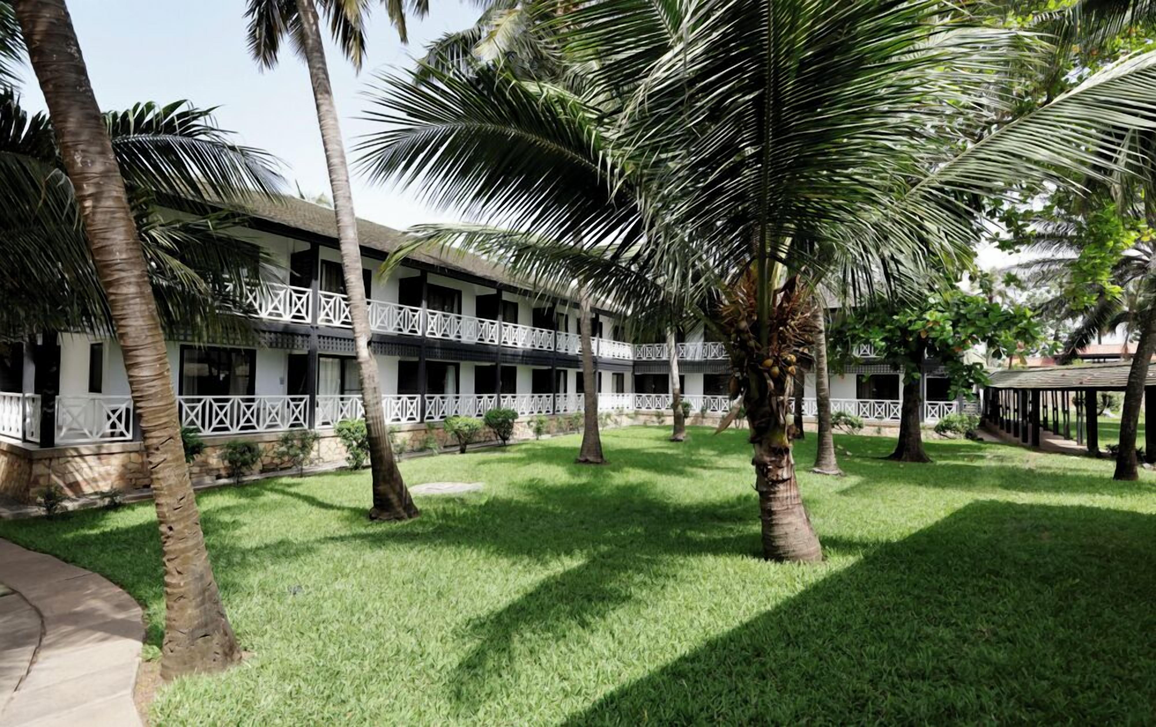 A hotel with a two-story structure, white balconies with geometric patterns, and a well-maintained lawn with palm trees.