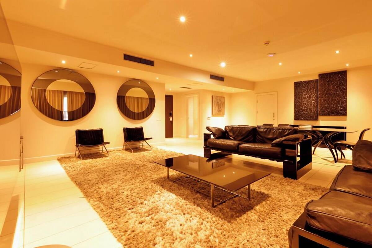 A modern living room with a leather sofa, a glass coffee table, and a fluffy rug. There are two chairs, a dining table, and a large mirror on the wall.
