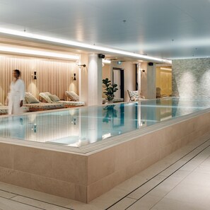 Zona interior de piscina moderna con barandilla de cristal, sillas de descanso y una persona caminando.