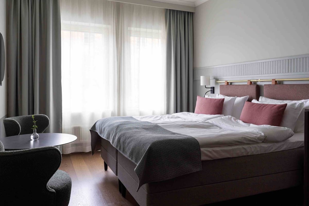 Une chambre d’hôtel avec un lit, une chaise, une table et une fenêtre avec des rideaux.