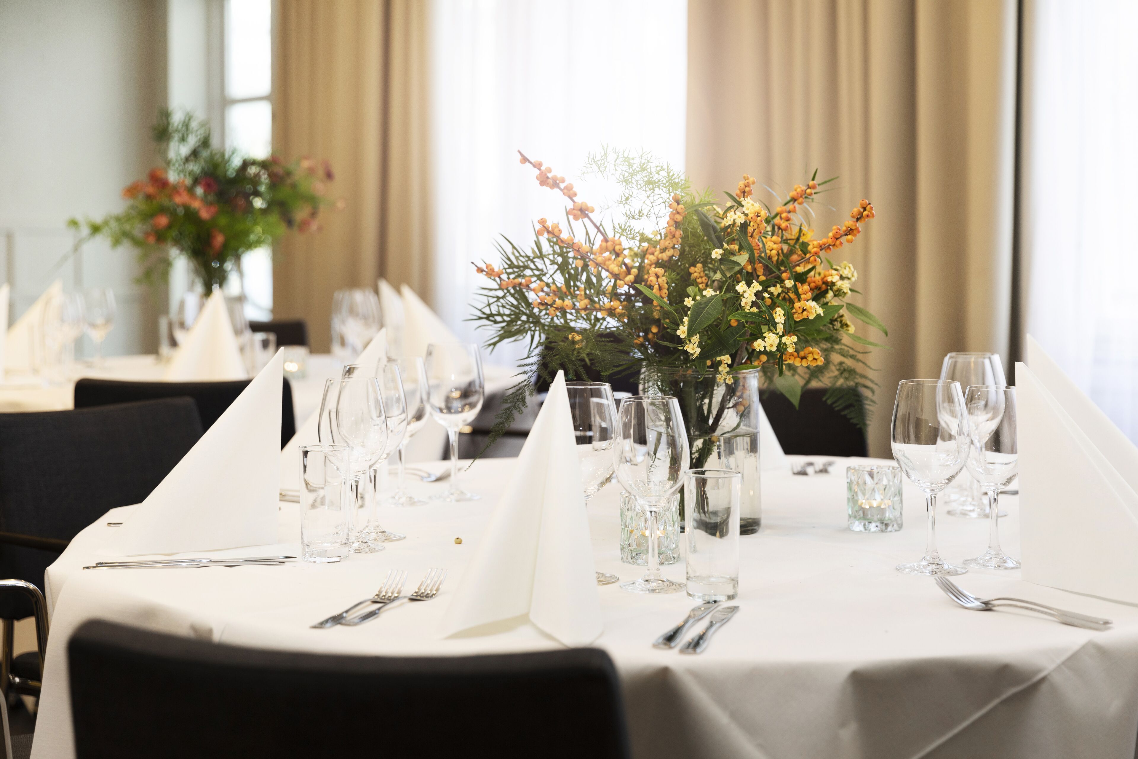 A dining table set with white tablecloths, glassware, and a centerpiece of yellow flowers and greenery.