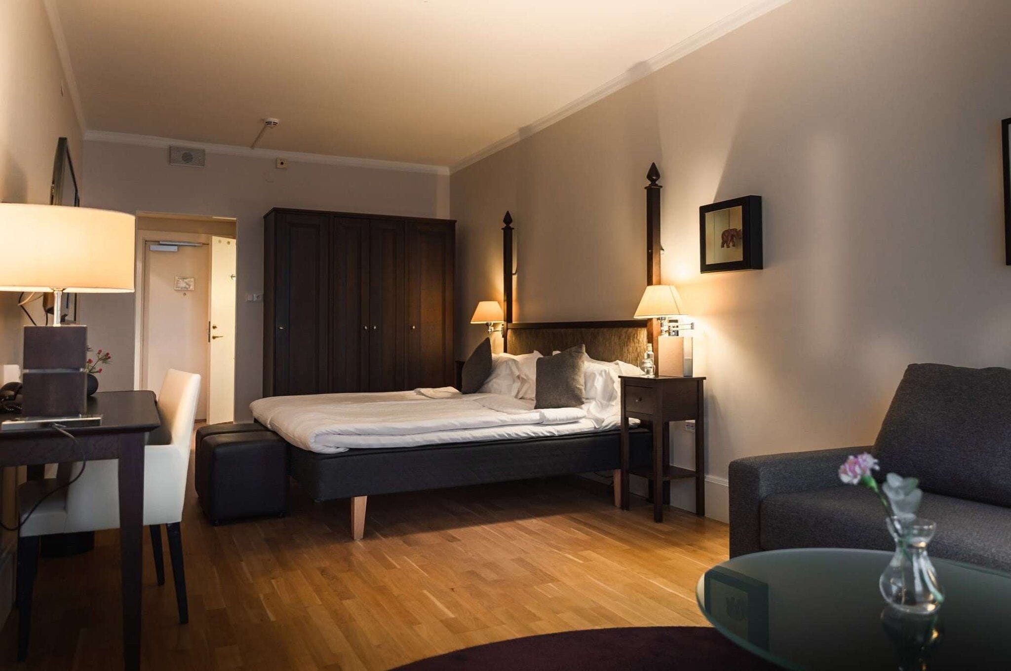 A hotel room with a bed, a desk, a chair, a sofa, and a small table with a vase.