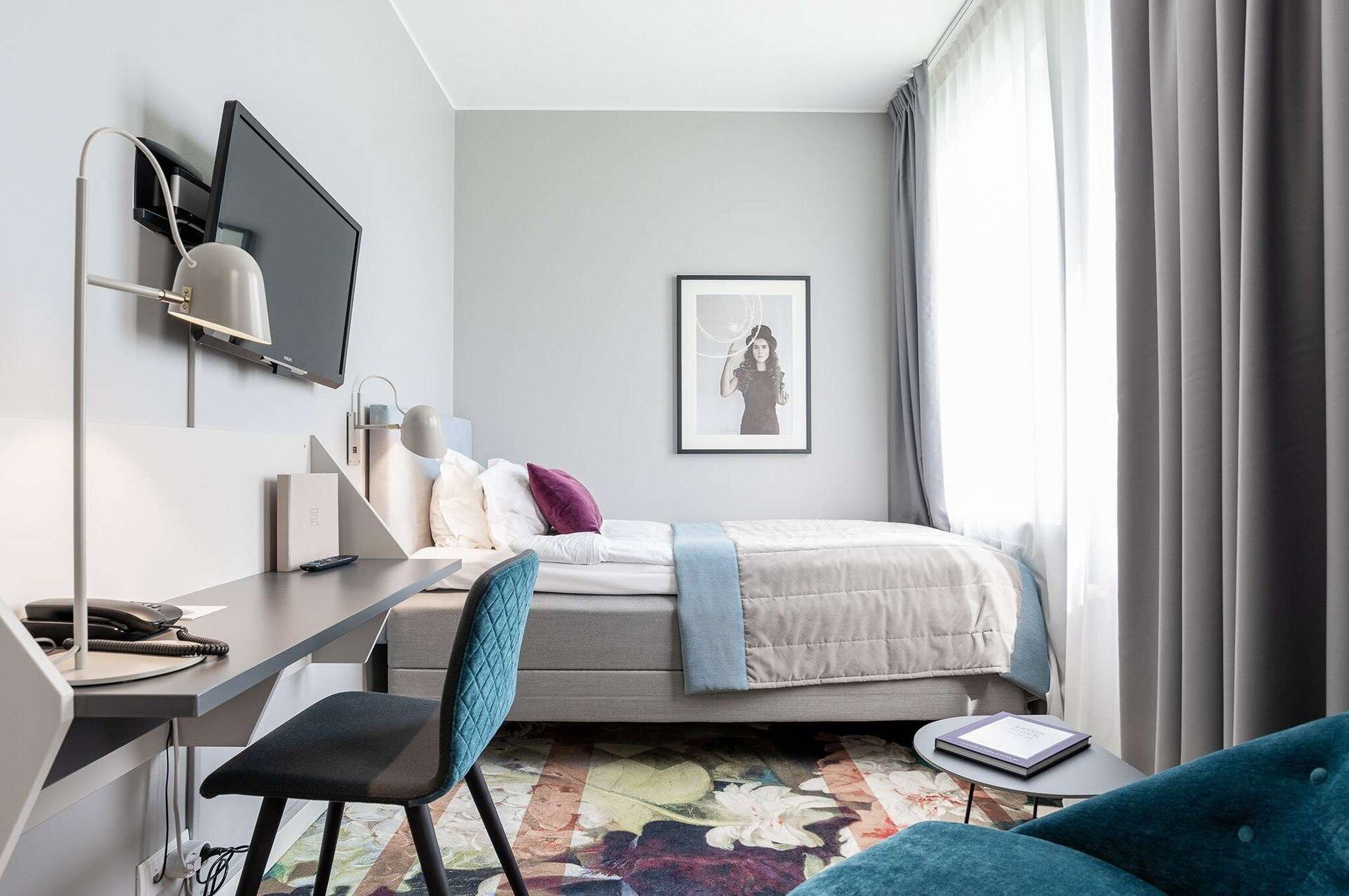 A modern hotel room with a bed, desk, chair, and a large window with curtains.