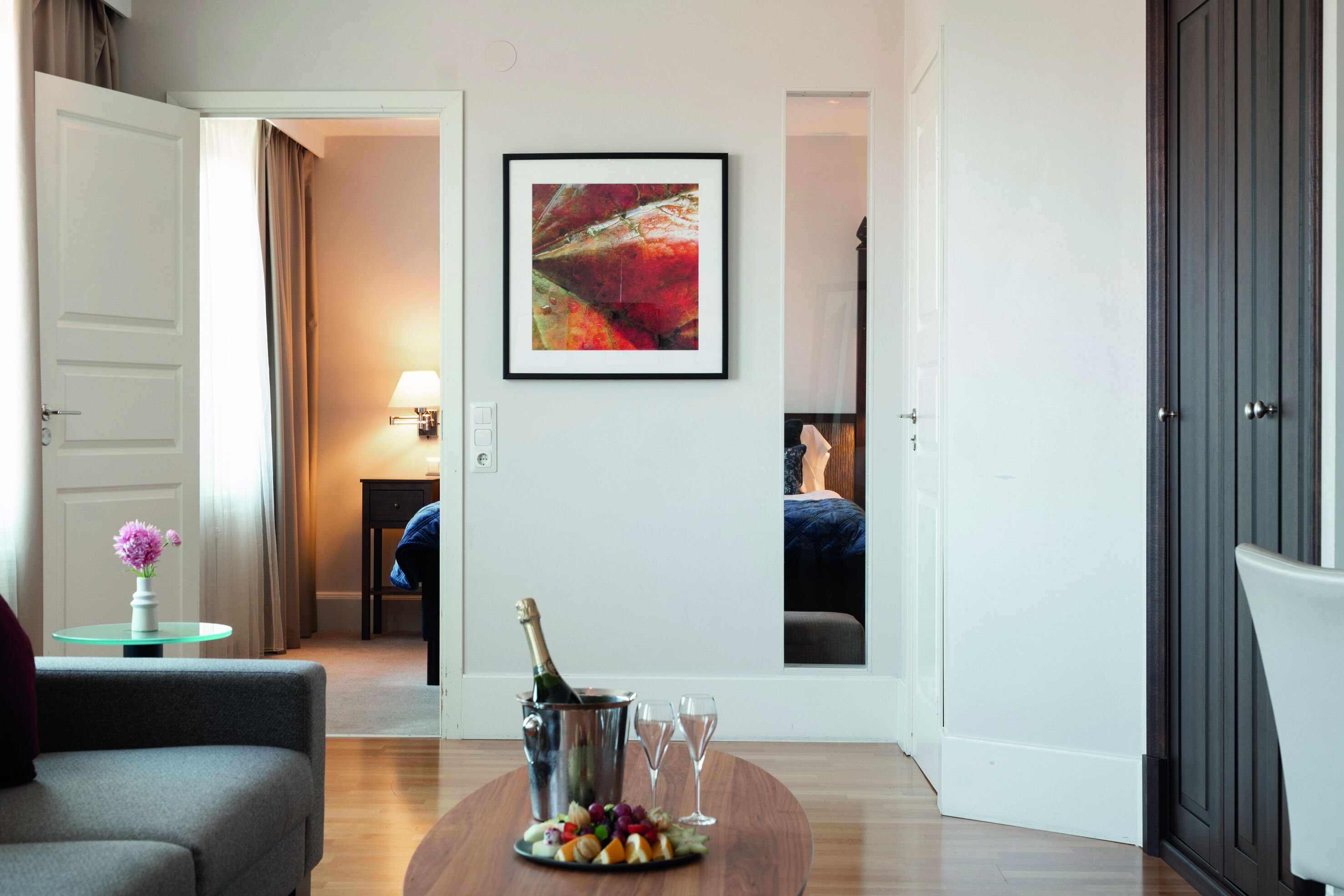 Quarto de hotel com piso de madeira, sofá cinza, pequena mesa com um espeto de frutas e champanhe, pintura abstrata emoldurada e guarda-roupa de madeira escura.