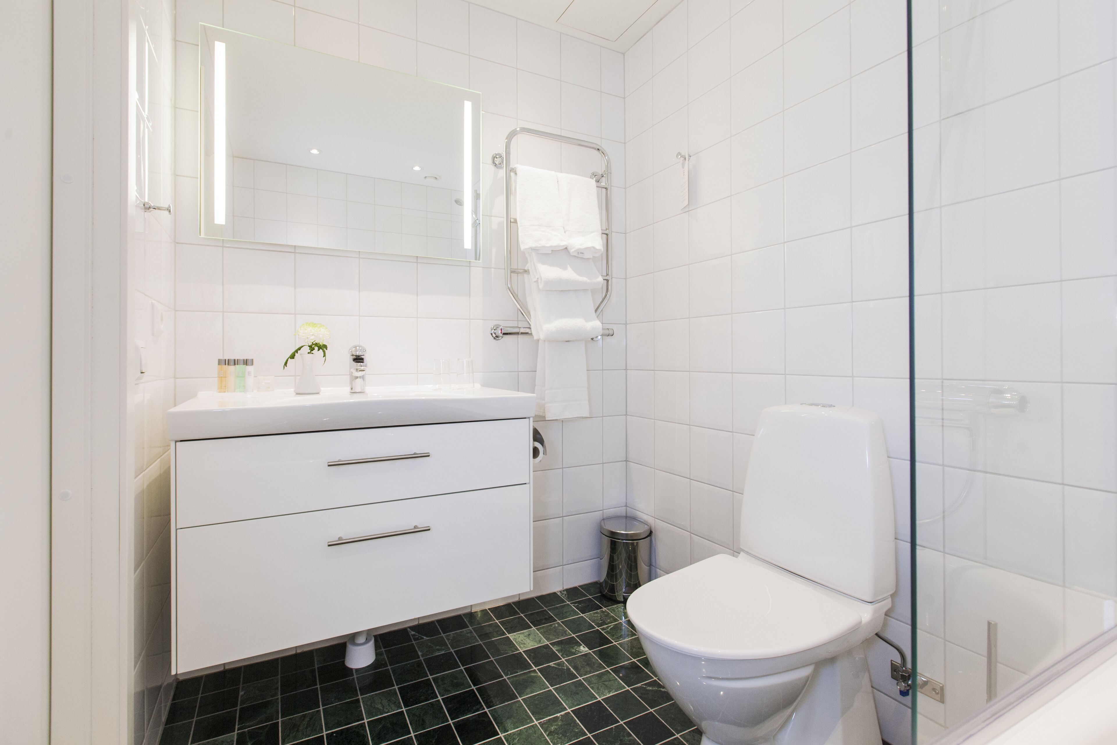 A modern bathroom with white tiles, a white toilet, a glass shower enclosure, and a vanity with a mirror and two drawers.