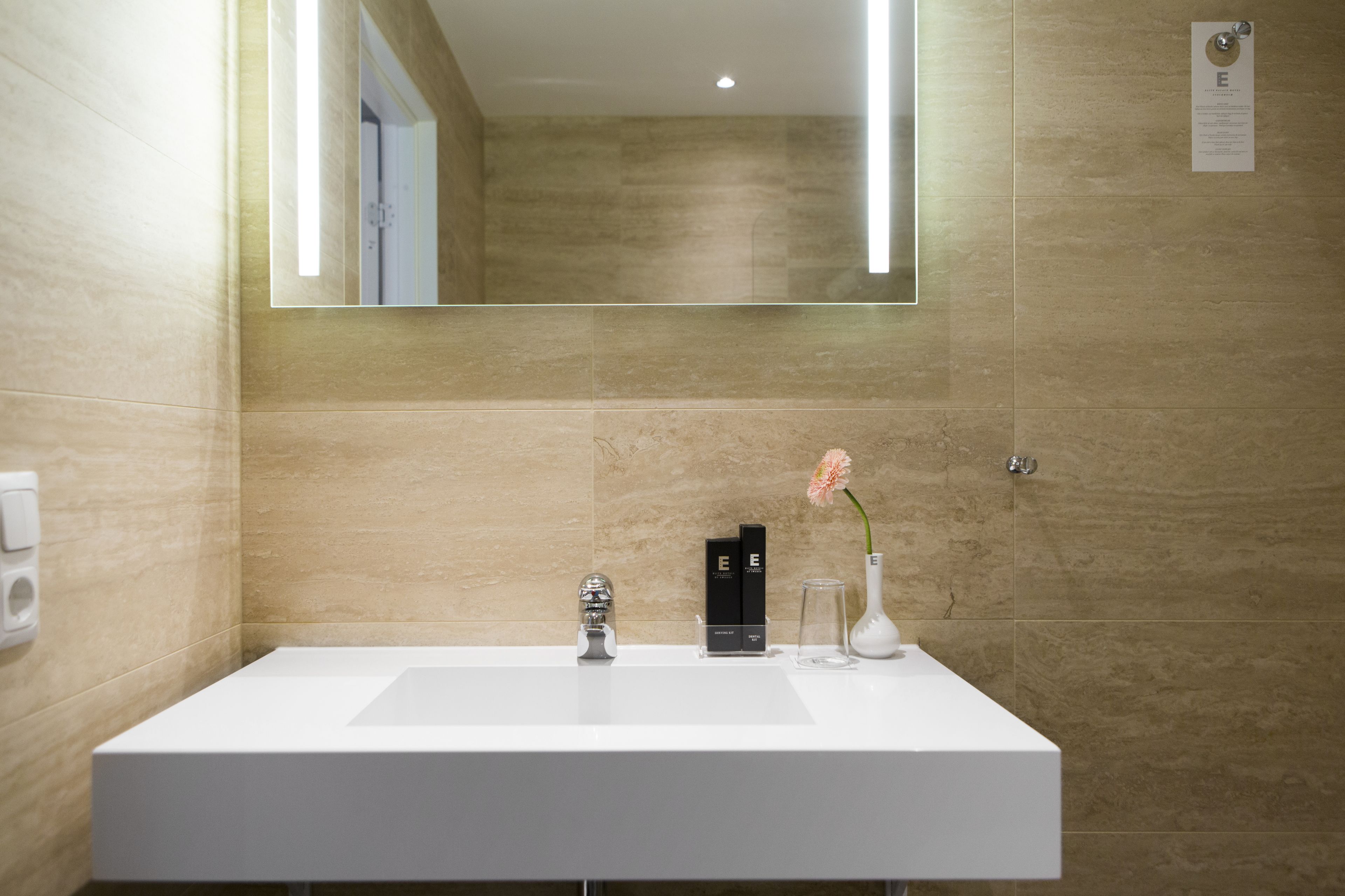 Une salle de bain moderne avec un lavabo blanc, un grand miroir et des murs recouverts de carreaux beiges.