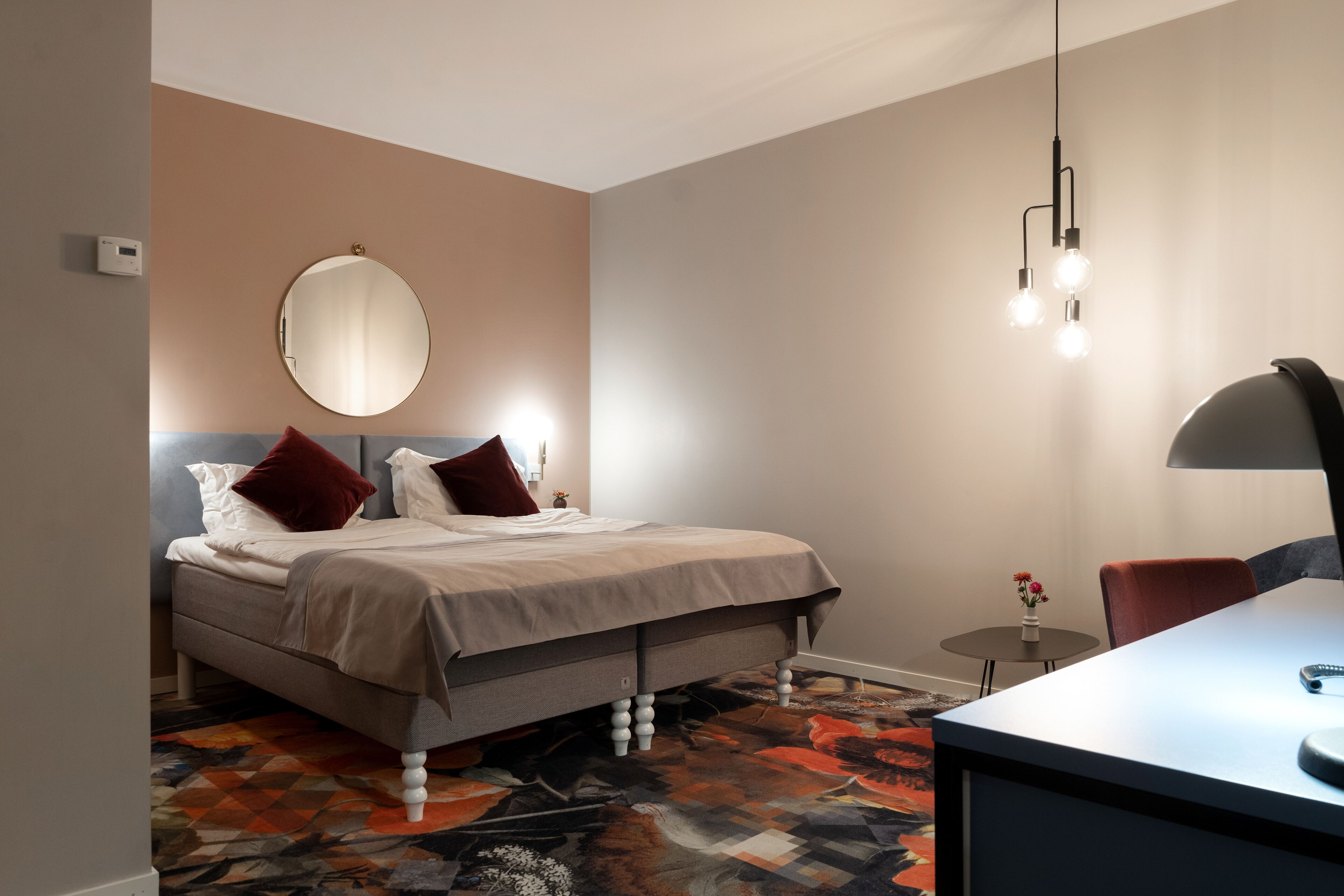 A hotel room with a bed, bedside table, lamp, and a small round table with flowers.