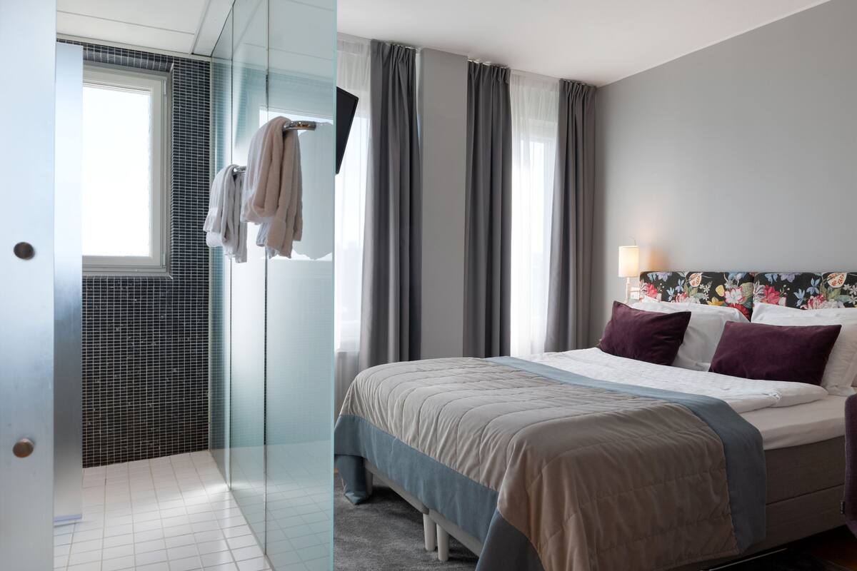 Une chambre d’hôtel moderne dotée d’une douche en verre, d’un lit avec une tête de lit à motifs floraux et offrant une vue sur le paysage urbain.