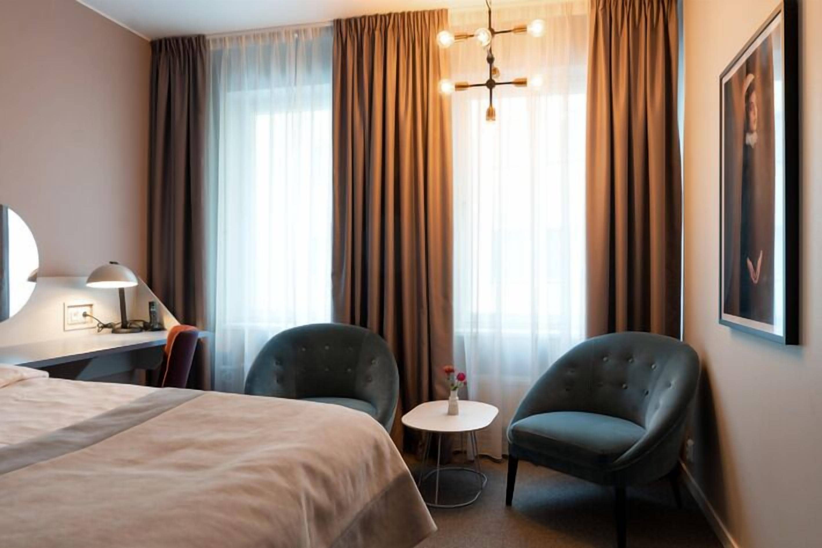 Ett hotellrum med en säng, två fåtöljer, ett litet runt bord med en vas, ett skrivbord med en lampa och ett stort fönster med gardiner.