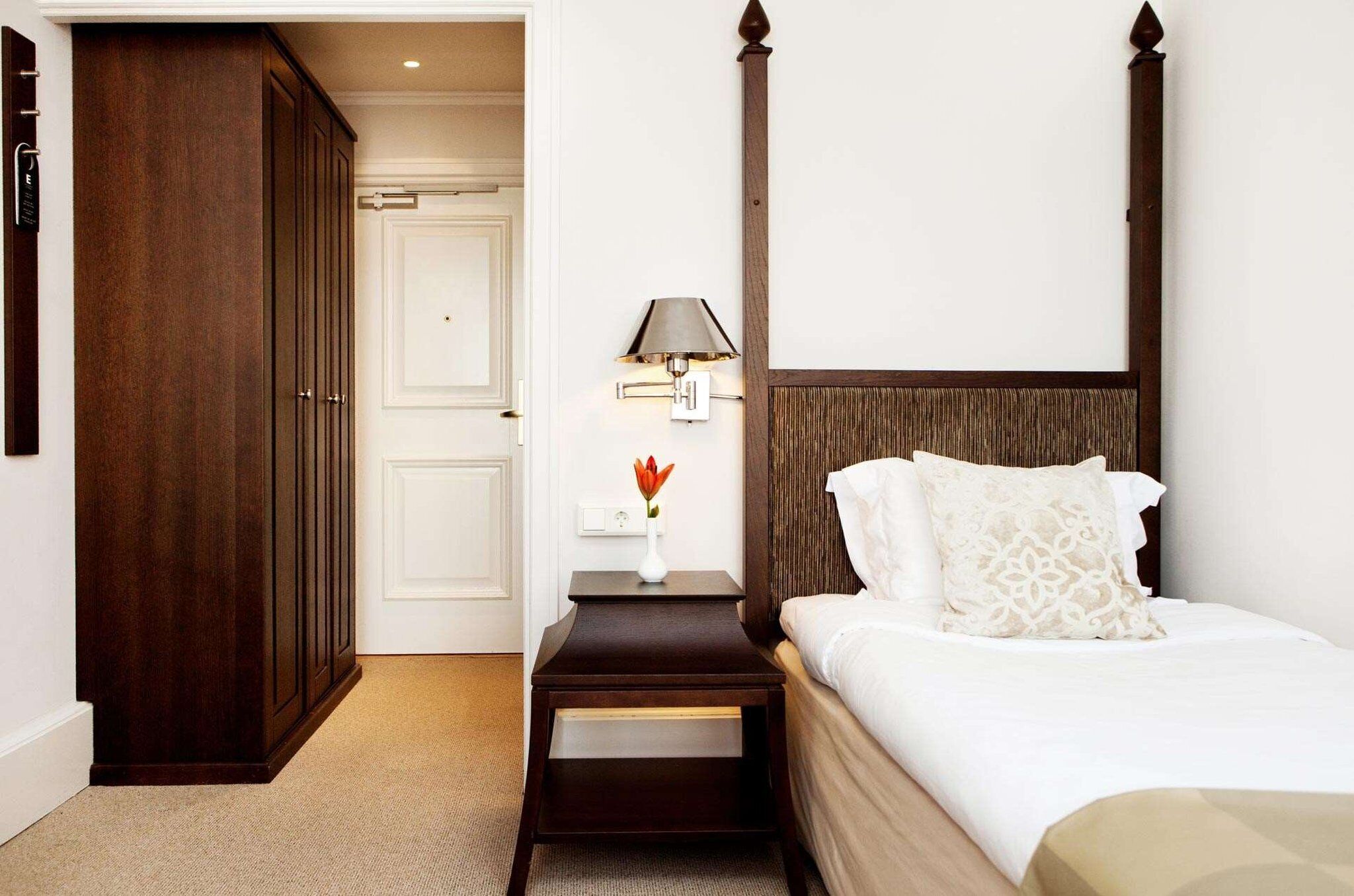 Une chambre d’hôtel avec une grande armoire en bois, une table de chevet avec une lampe et un lit avec du linge de lit blanc.