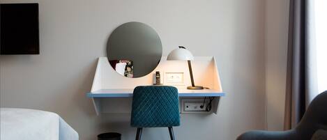 A hotel room with a desk, a chair, a round mirror, a lamp, and a TV mounted on the wall.