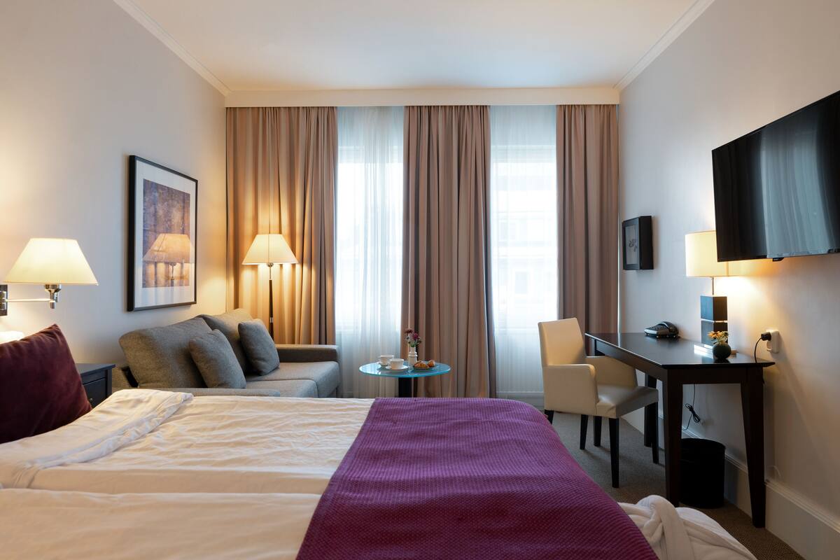 Une chambre d’hôtel avec un grand lit, un bureau avec une chaise, une petite table avec une tasse et un vase, et un téléviseur fixé au mur.