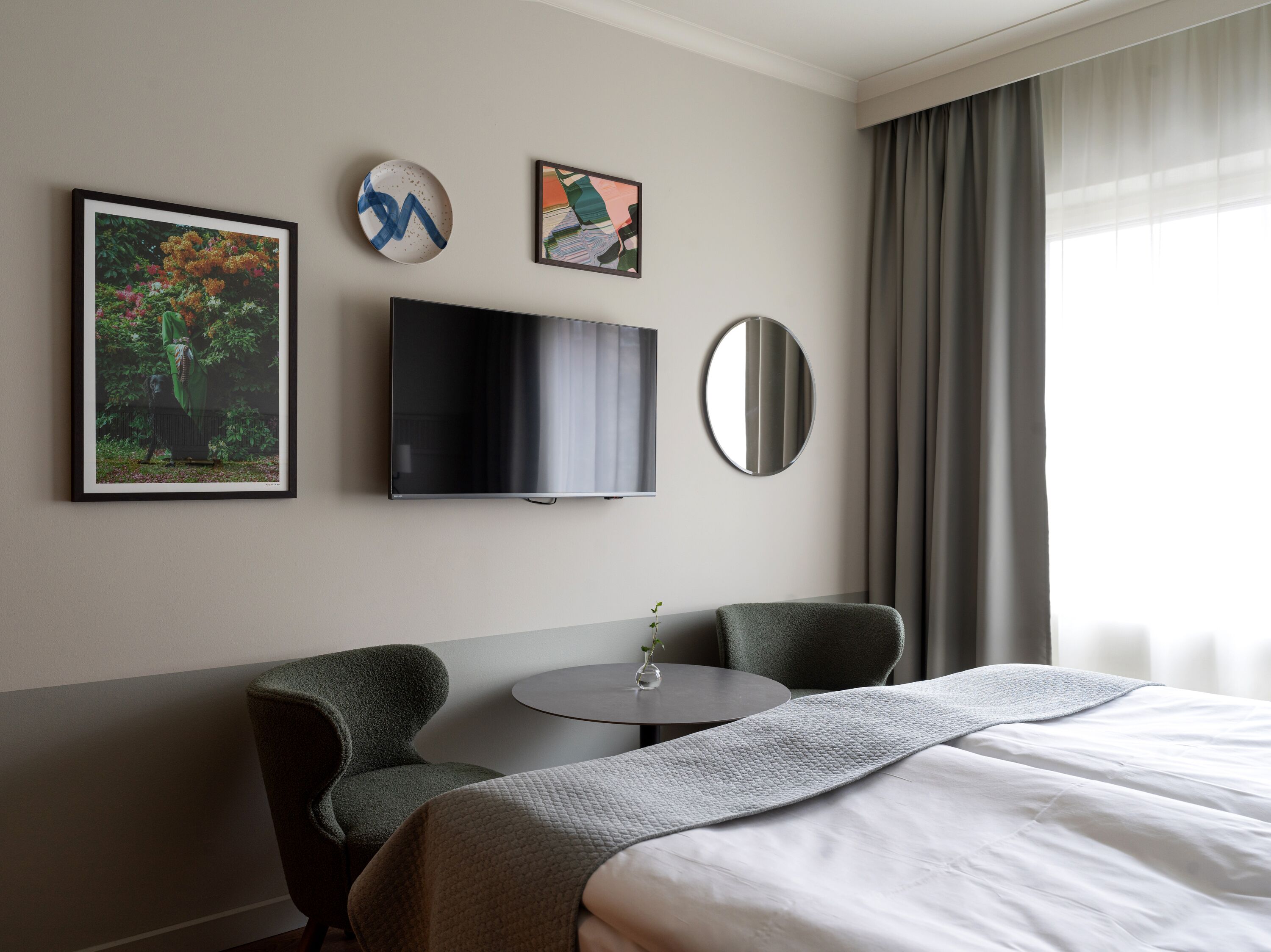 Habitación de hotel con cama, dos sillas, televisión, espejo redondo y cuadros en la pared.