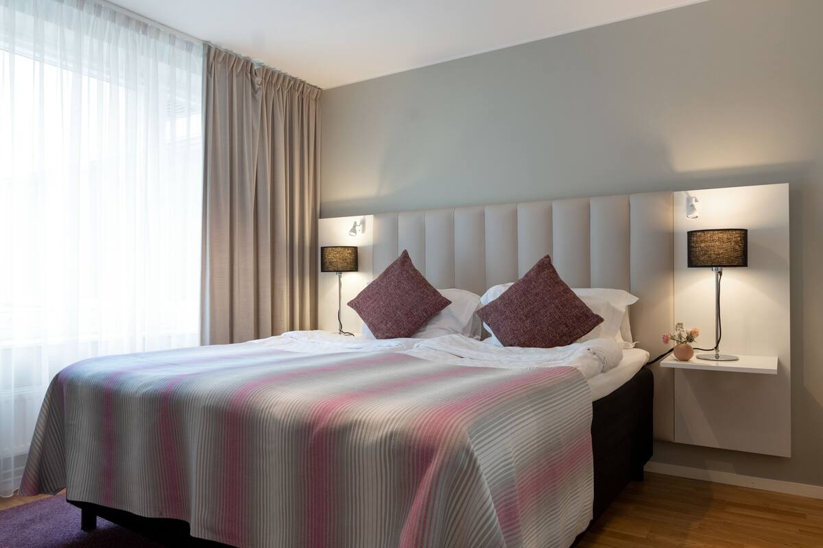 Un lit bien fait, recouvert d’une couvre-lit à rayures, avec deux lampes de chevet et une table de nuit ornée d’un vase de fleurs.