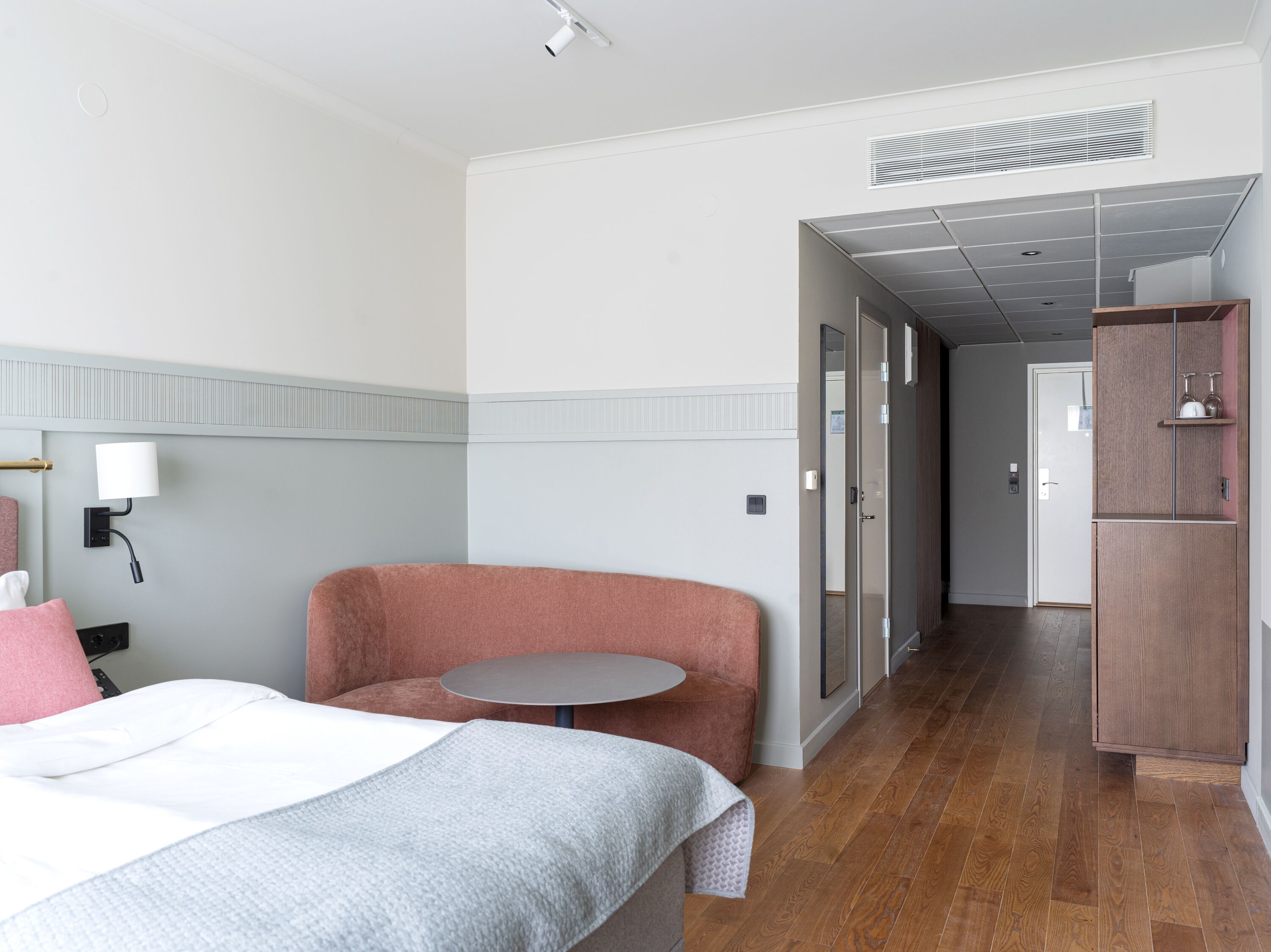 A hotel room with a bed, a pink armchair, a small round table, a wall-mounted lamp, and a wardrobe.