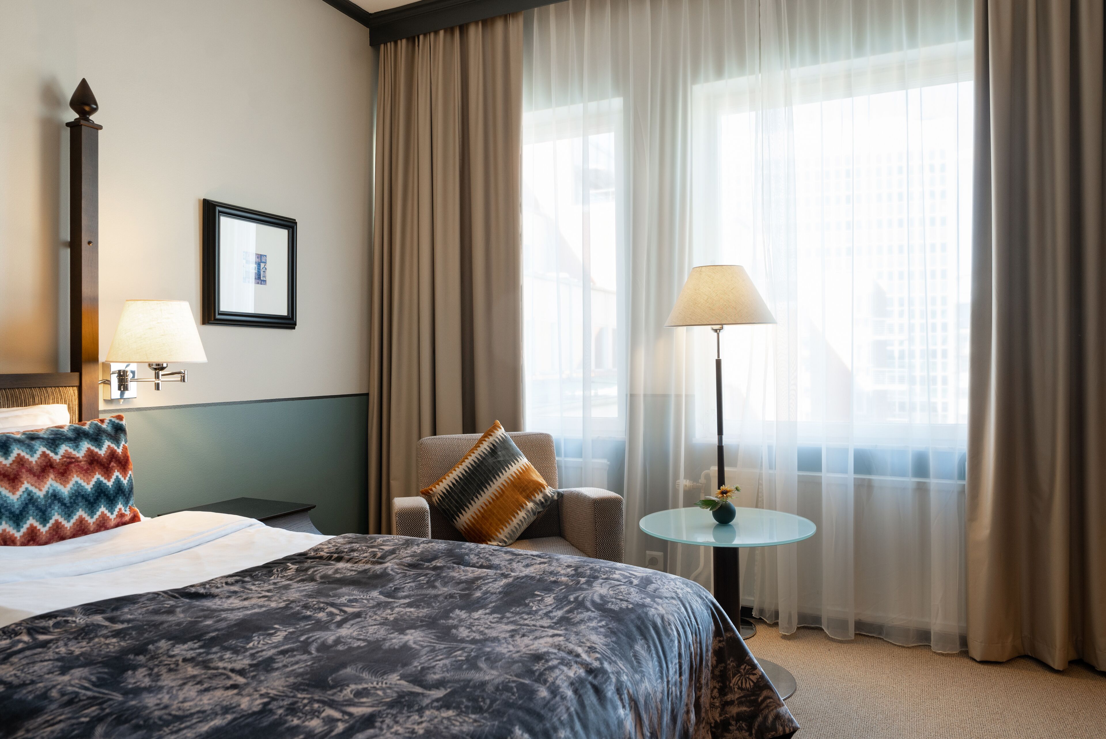 Habitación de hotel con una cama grande, una silla, una mesita con lámpara y una ventana con cortinas.