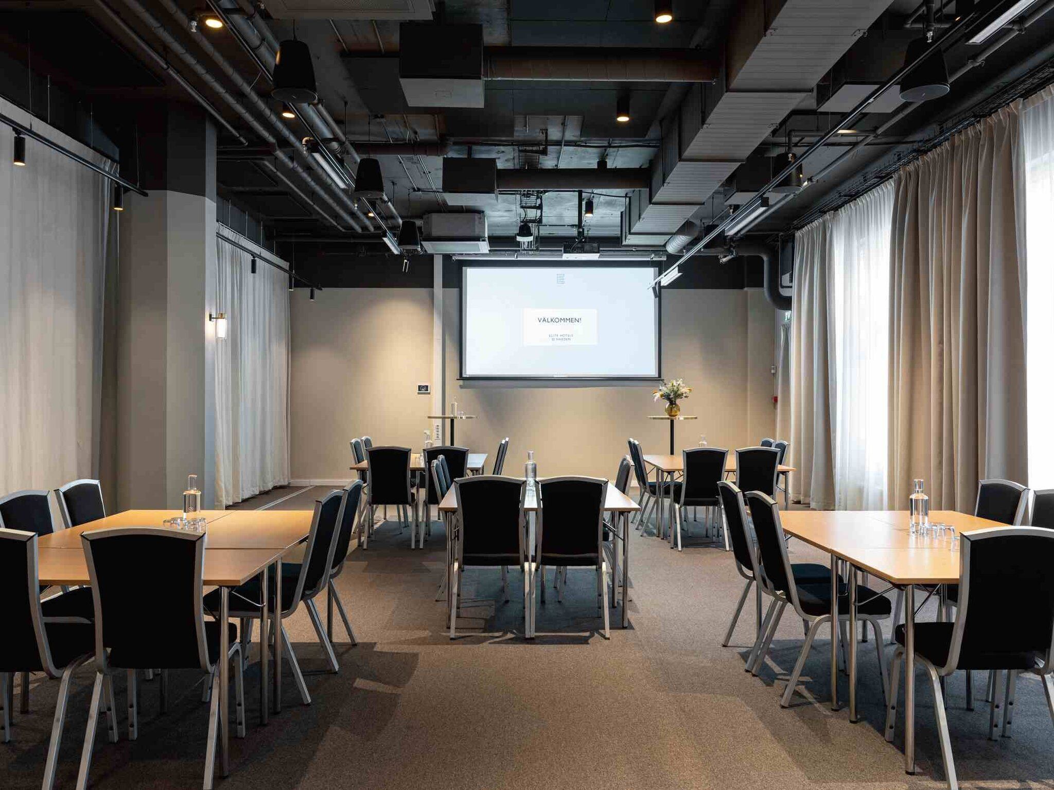 A conference room with a large screen displaying 'Valkommen' and a projector. The room has a modern design with black chairs and tables, and a carpeted floor.