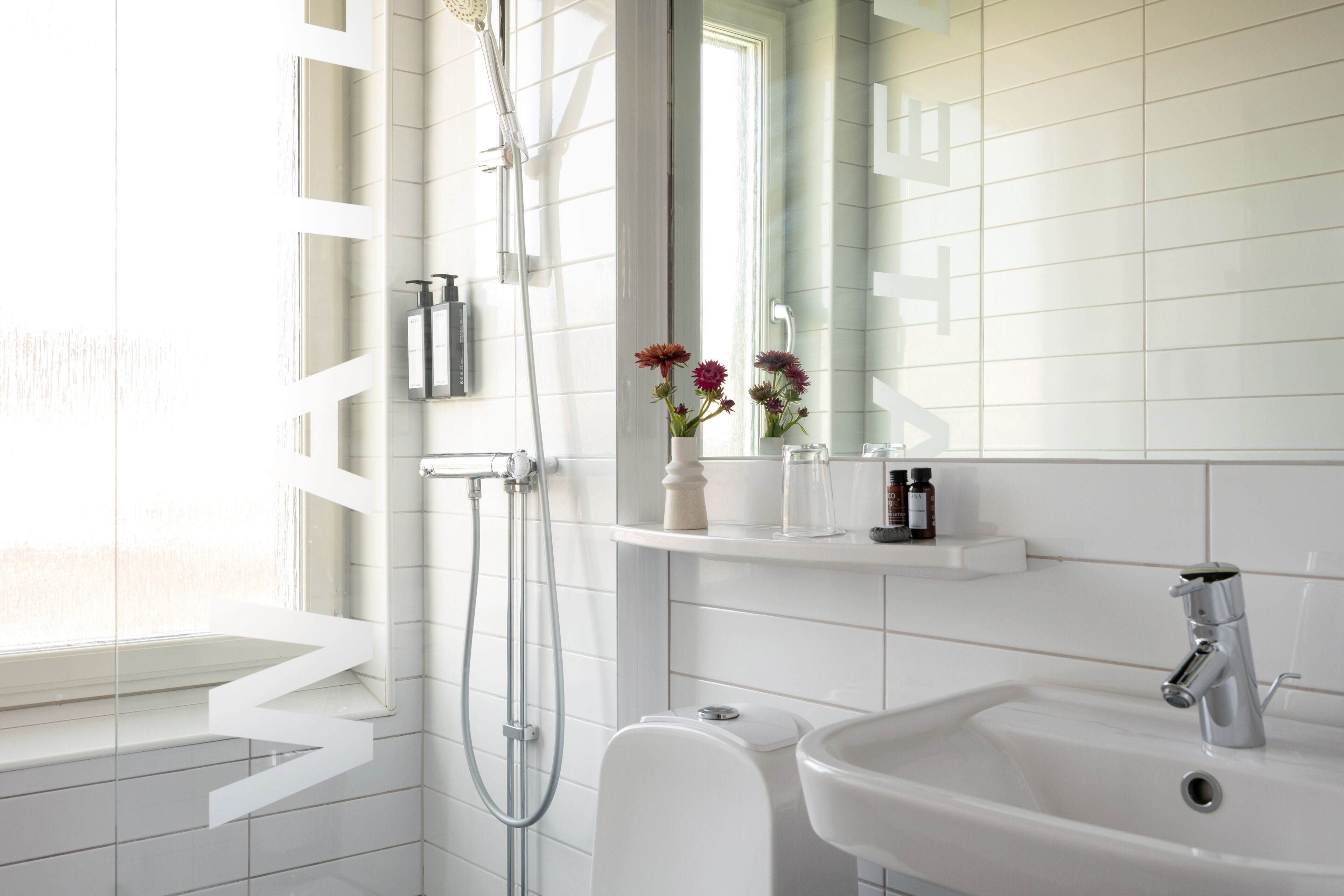 Ein modernes Badezimmer mit einem weißen Waschbecken, einem Spiegel und einer Dusche. Auf dem Waschbecken stehen Blumenarrangements und ein Fenster sorgt für natürliches Licht.