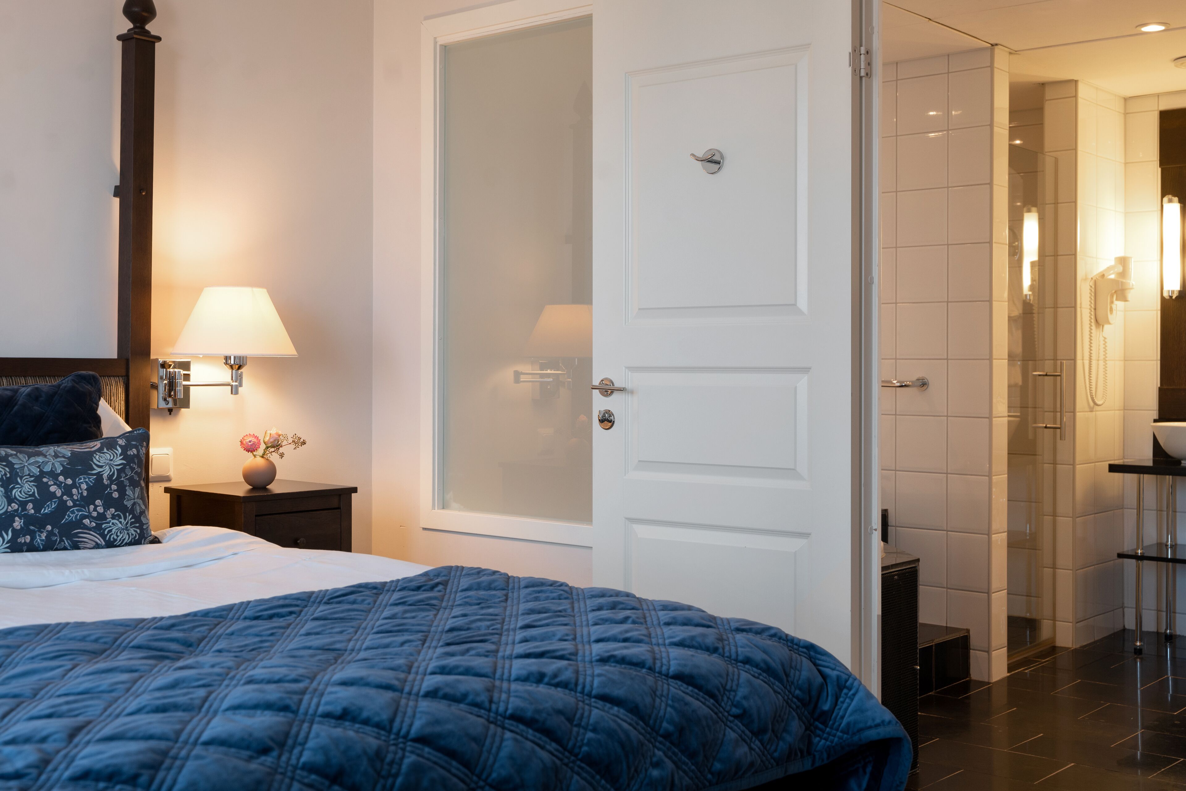 Ein Hotelzimmer mit einem Bett, einem Nachttisch mit Lampe, einer Tür zu einem Badezimmer und einer Duschkabine.