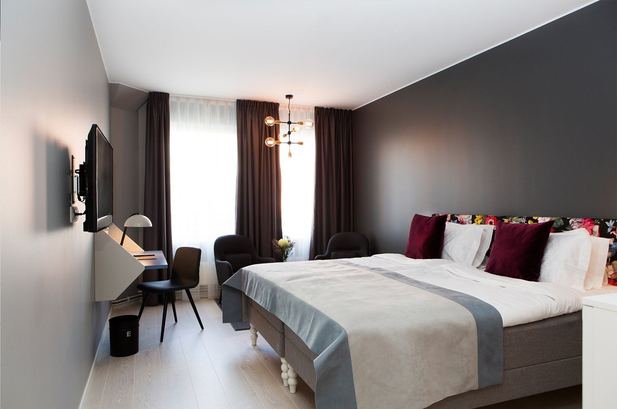 A modern hotel room with a large bed, a desk with a chair, a TV, and a window with curtains.