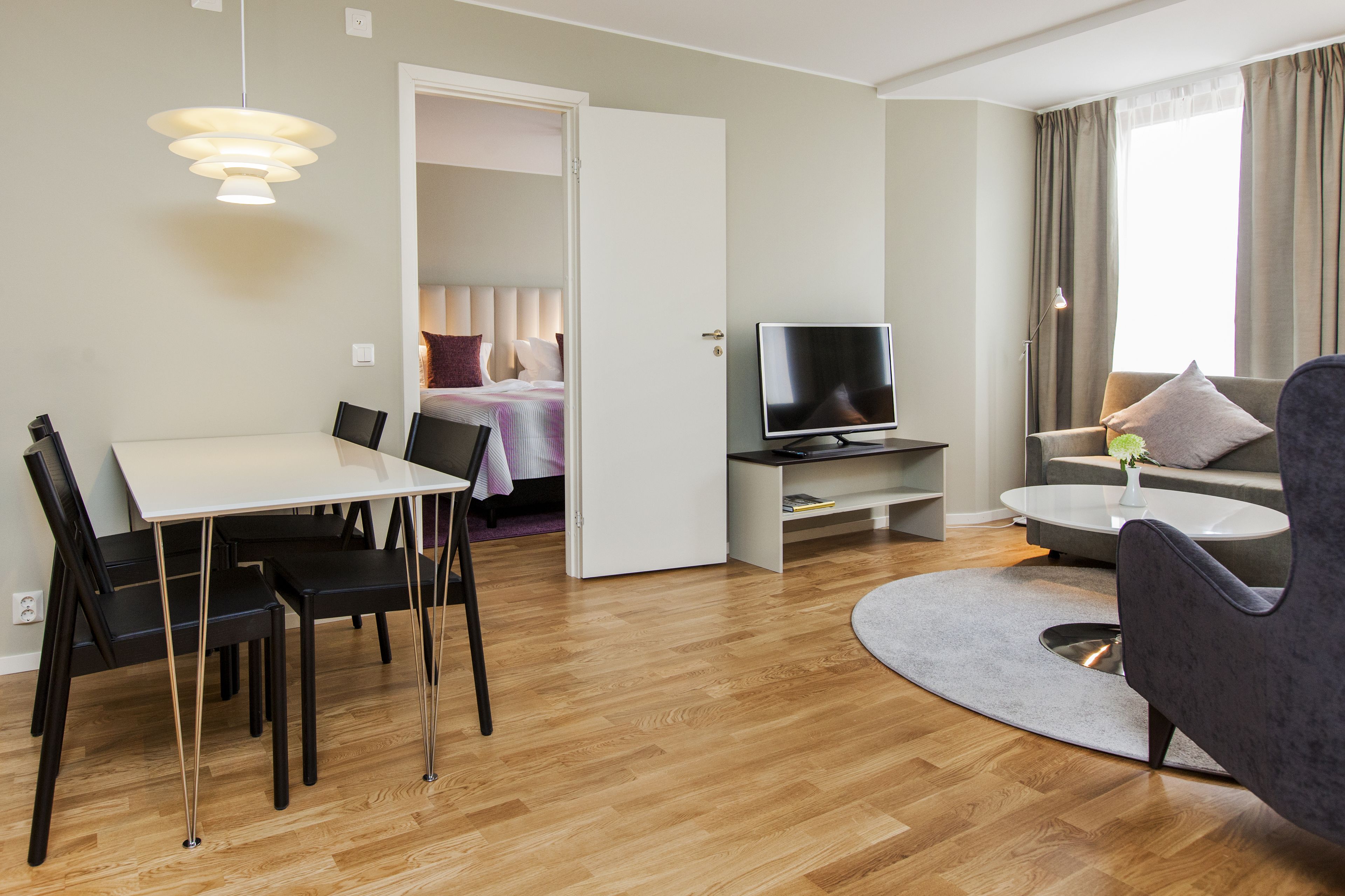 Et moderne hotelværelse med spiseplads, opholdsstue med sofa og fjernsyn, samt et soveværelse, der kan anes gennem en åben dør.