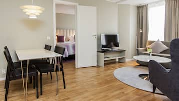 Et moderne hotelværelse med spiseplads, opholdsstue med sofa og fjernsyn, samt et soveværelse, der kan anes gennem en åben dør.