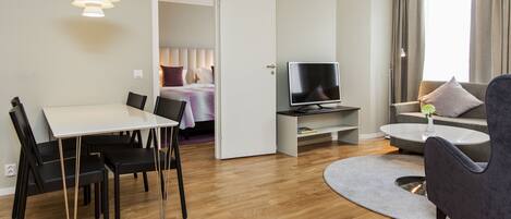 A modern hotel room with a dining area, a living space with a sofa and TV, and a bedroom visible through an open door.
