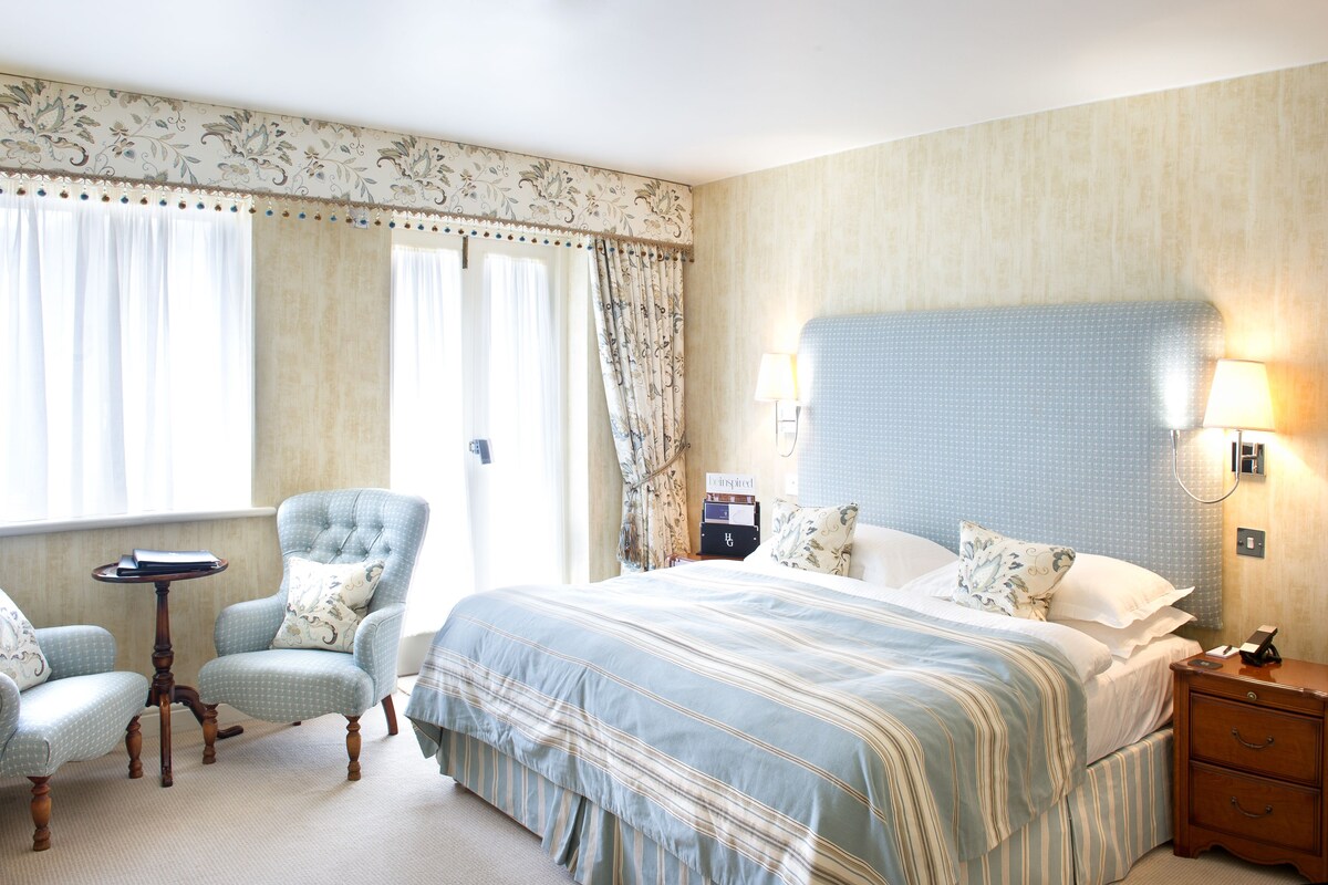 A hotel room with a large bed, two armchairs, a small table, and a nightstand with a lamp.
