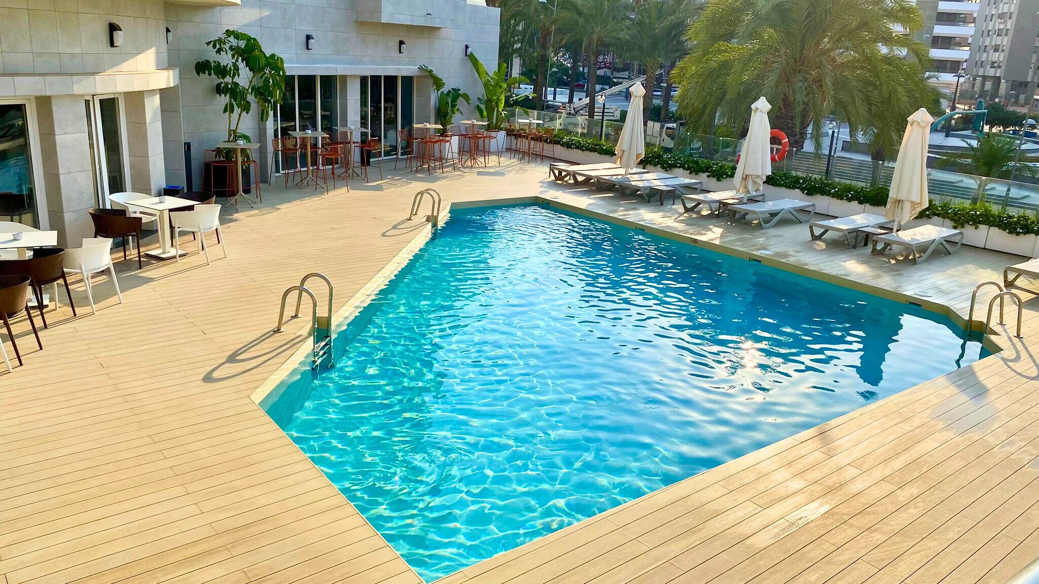 A rooftop pool with wooden decking, lounge chairs, and tables.