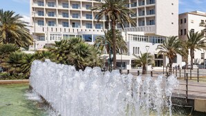 Un hôtel avec une fontaine d’eau devant, des palmiers et un ciel dégagé.
