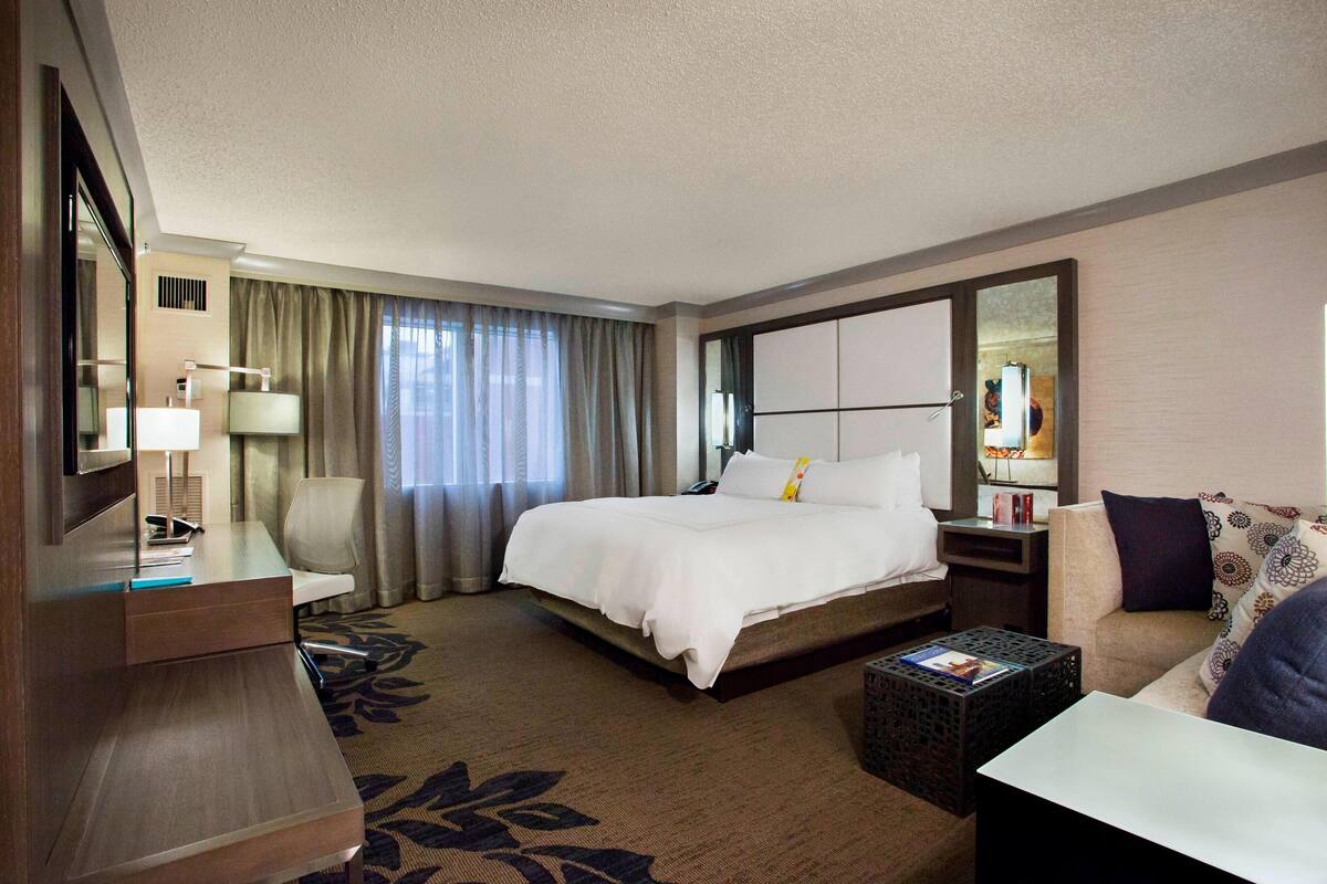 Quarto de hotel com uma cama grande, uma escrivaninha com luminária, uma cadeira, um sofá e uma mesa pequena.
