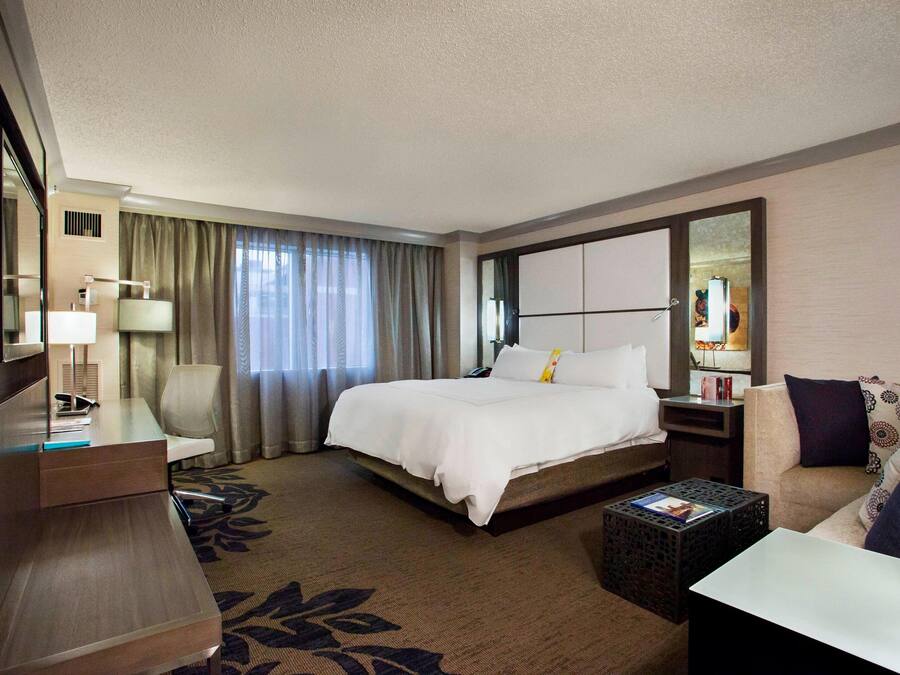 A hotel room with a large bed, a desk with a lamp, a chair, a sofa, and a small table.