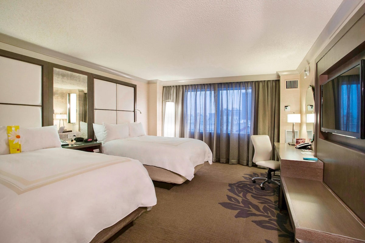 Quarto de hotel com duas camas, uma escrivaninha, uma cadeira, televisão e uma janela grande com cortinas.
