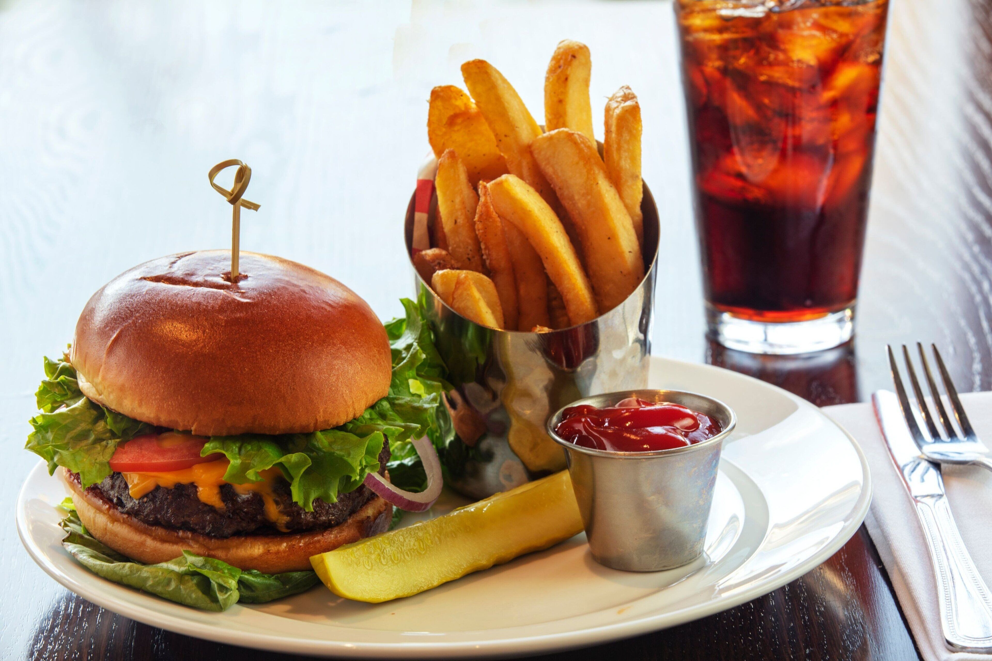 Un hamburger avec des frites et un cornichon sur une assiette, un verre de thé glacé et une fourchette posée sur une serviette.