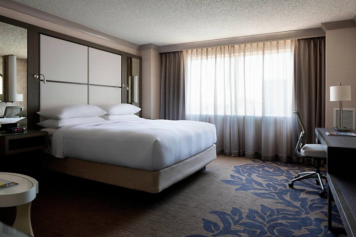 Quarto de hotel com uma cama grande, uma escrivaninha, uma cadeira, uma mesa de cabeceira e uma janela ampla com cortinas.