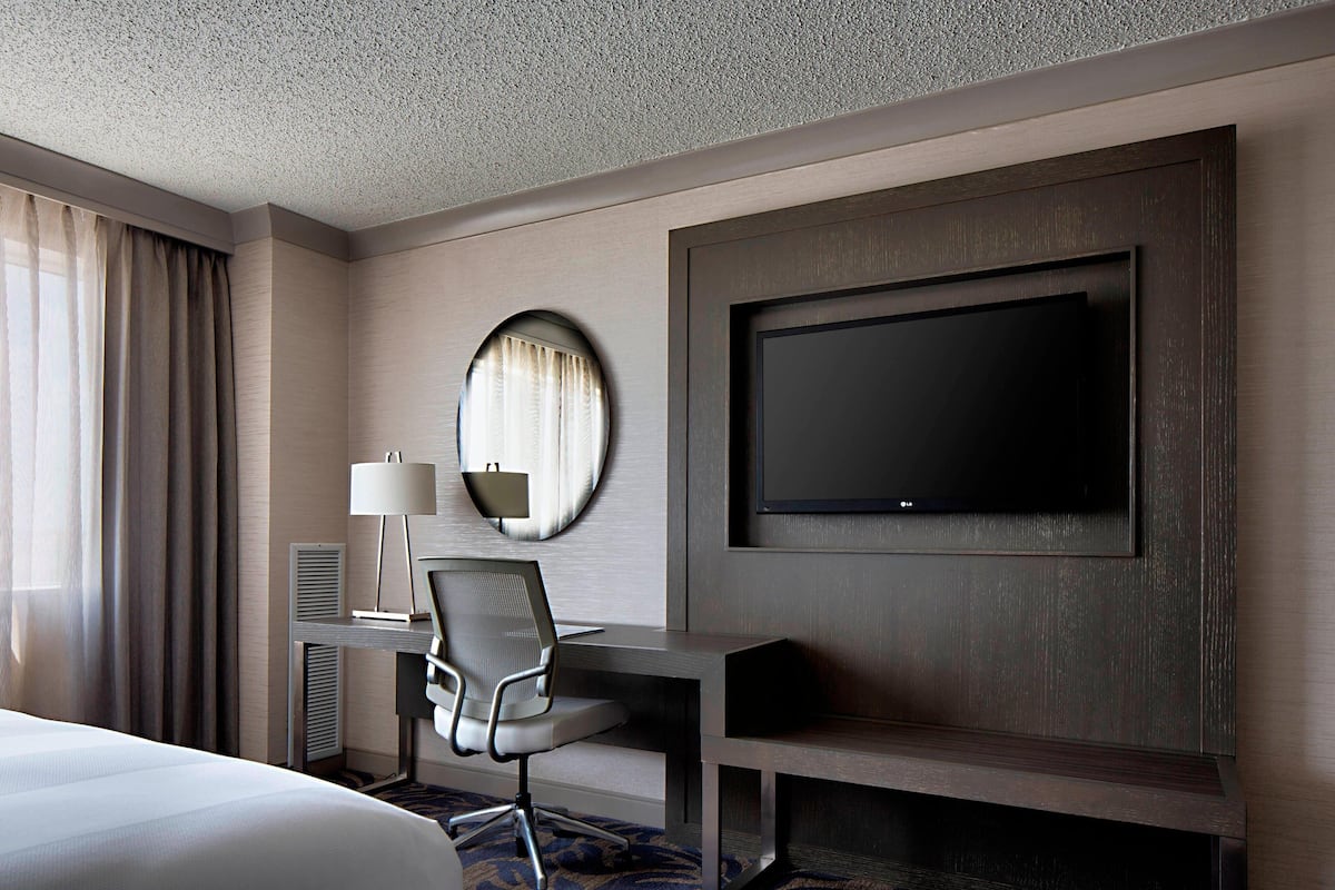 Quarto de hotel com uma cama grande, uma escrivaninha com cadeira, televisão de tela plana na parede, um espelho redondo e uma janela com cortinas.