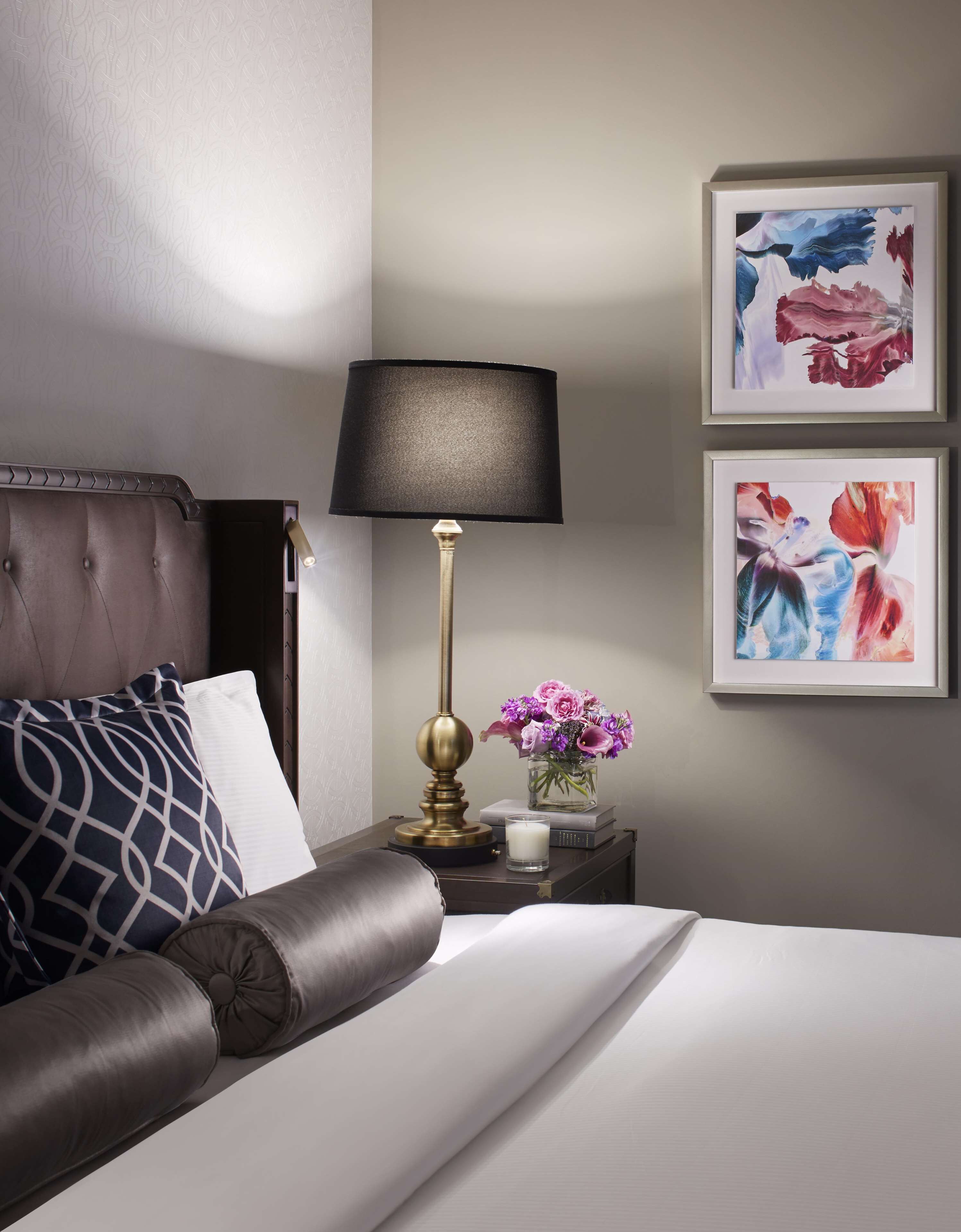 A neatly made bed with a dark headboard, a bedside table with a lamp, a vase with flowers, and two framed artworks on the wall.