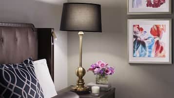 A neatly made bed with a dark headboard, a bedside table with a lamp, a vase with flowers, and two framed artworks on the wall.