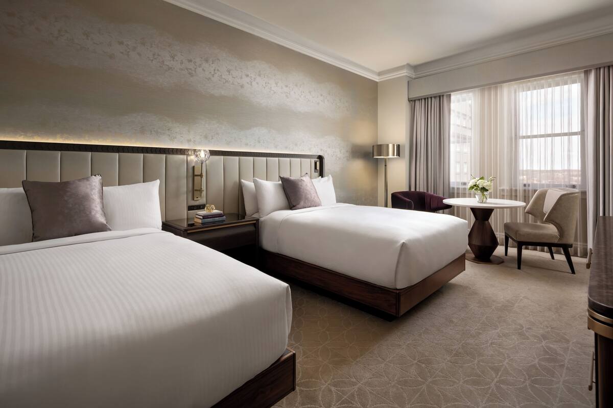 A hotel room with two beds, a bedside table with a lamp, a small round table with a vase of flowers, and a chair.