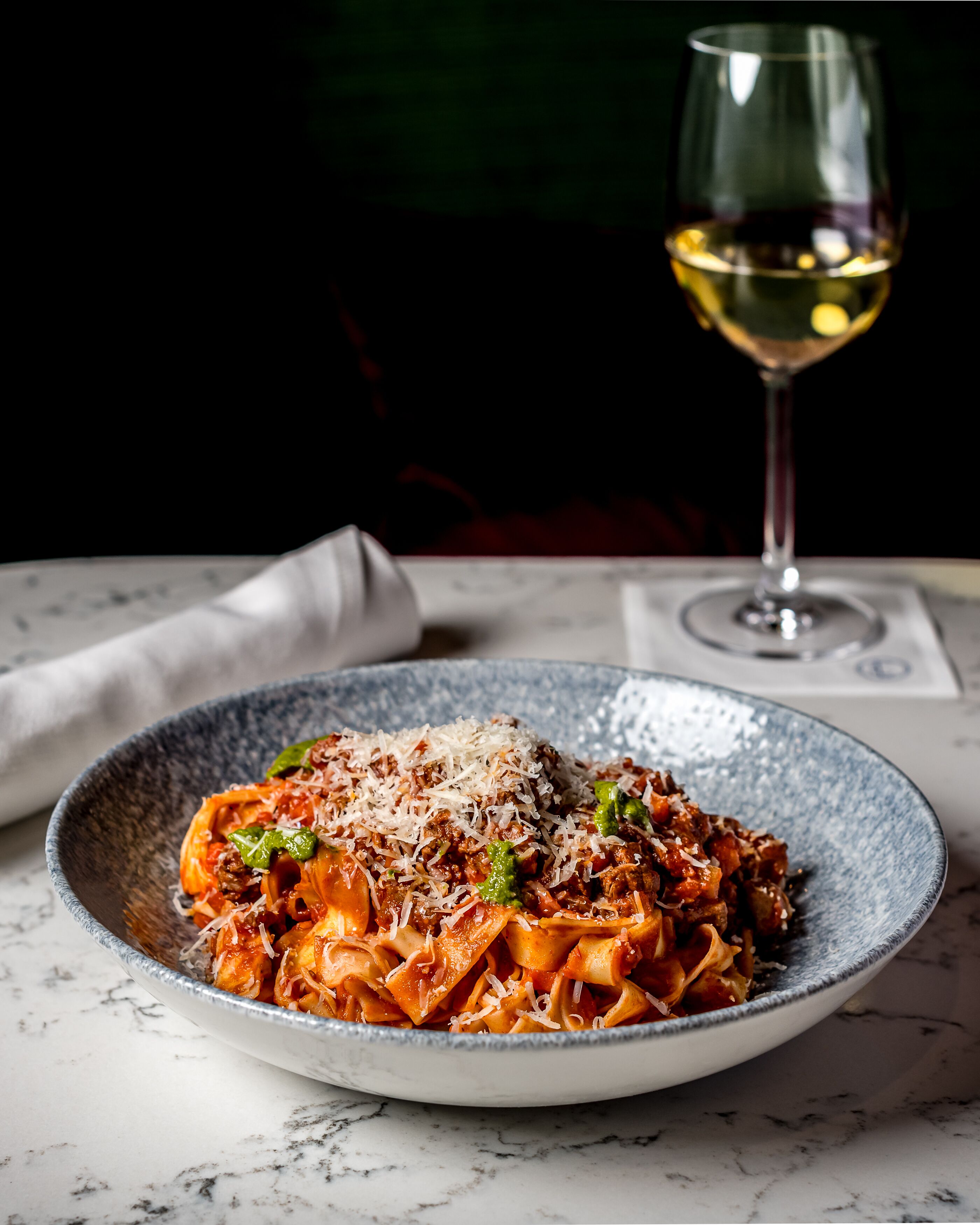A plate of pasta with meat and cheese, a glass of white wine, and a napkin on a marble table.