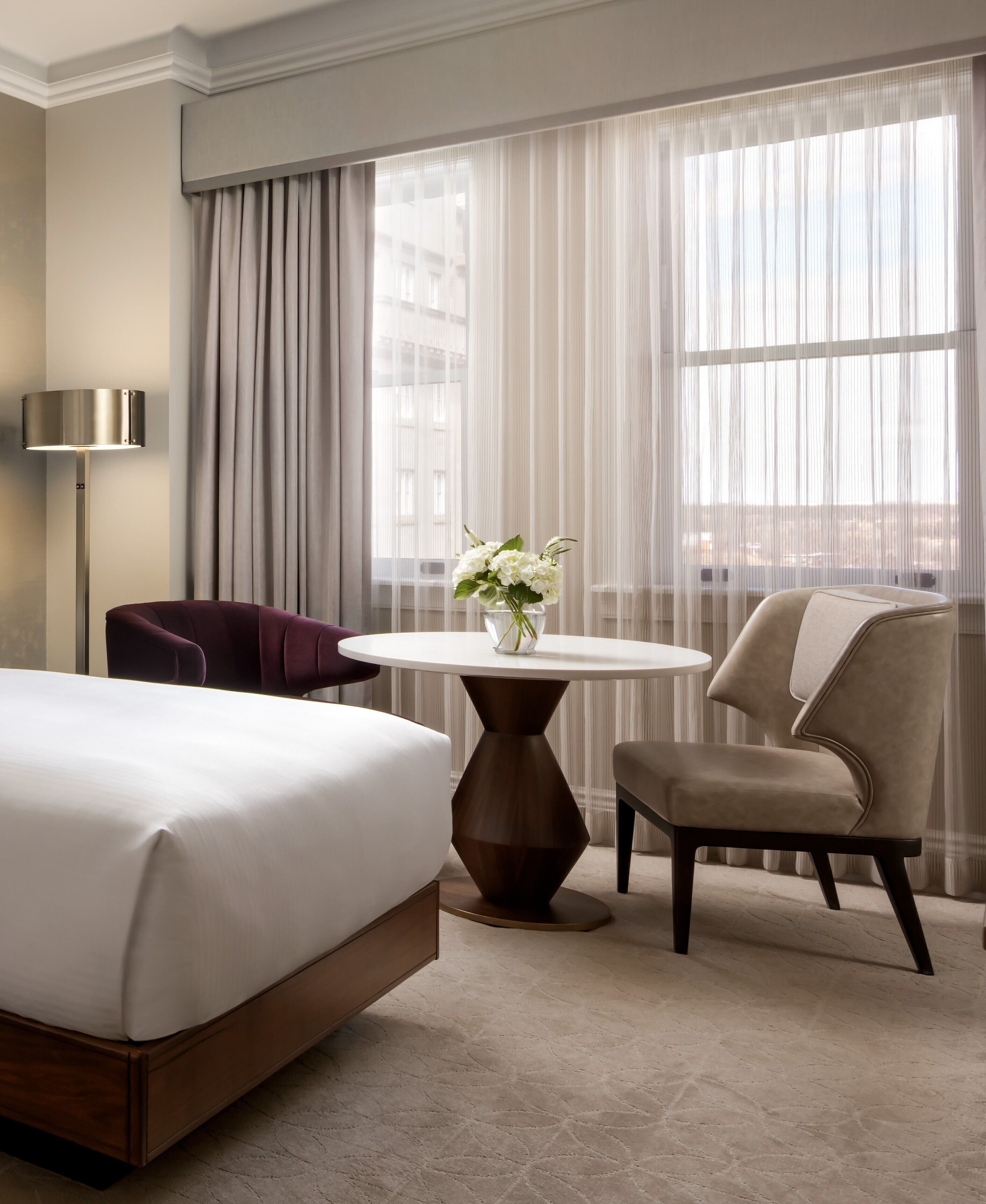 Une chambre d’hôtel avec un lit, une table ronde ornée d’un vase de fleurs, une chaise et une lampe.