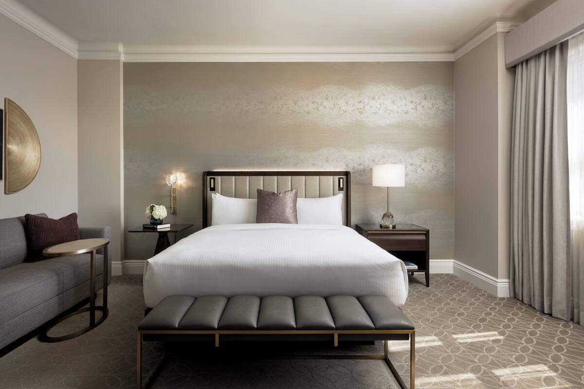A hotel room with a large bed, a bedside table with a lamp, a small round table, a sofa, and a bench.