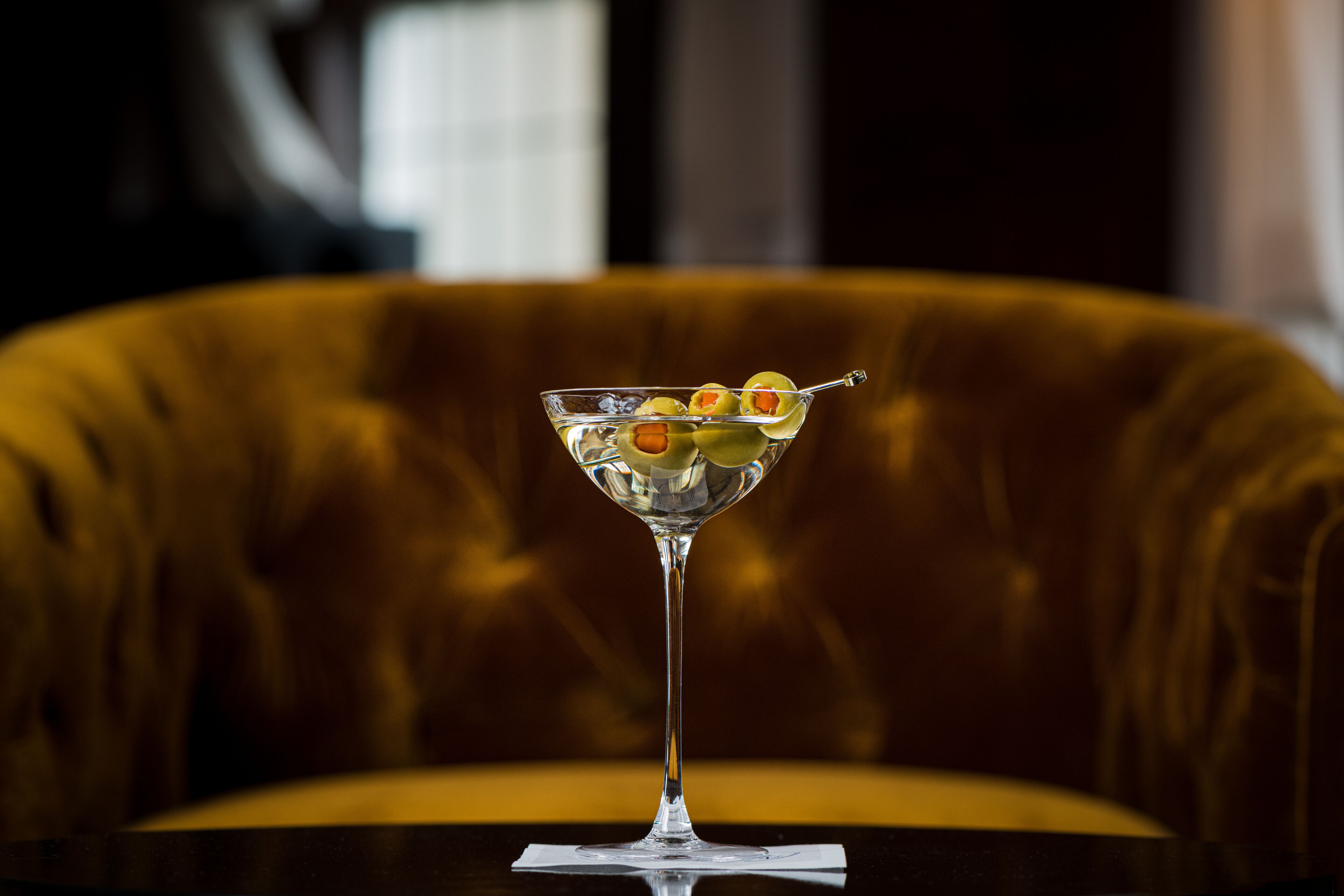 A martini glass with olives on a reflective surface, set against a plush, upholstered chair.