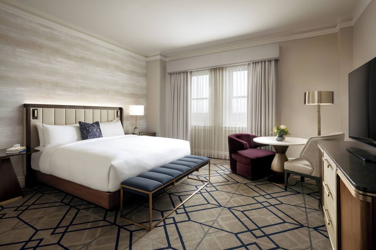 A hotel room with a large bed, a bench, a desk with a chair, a TV, and a window with curtains.