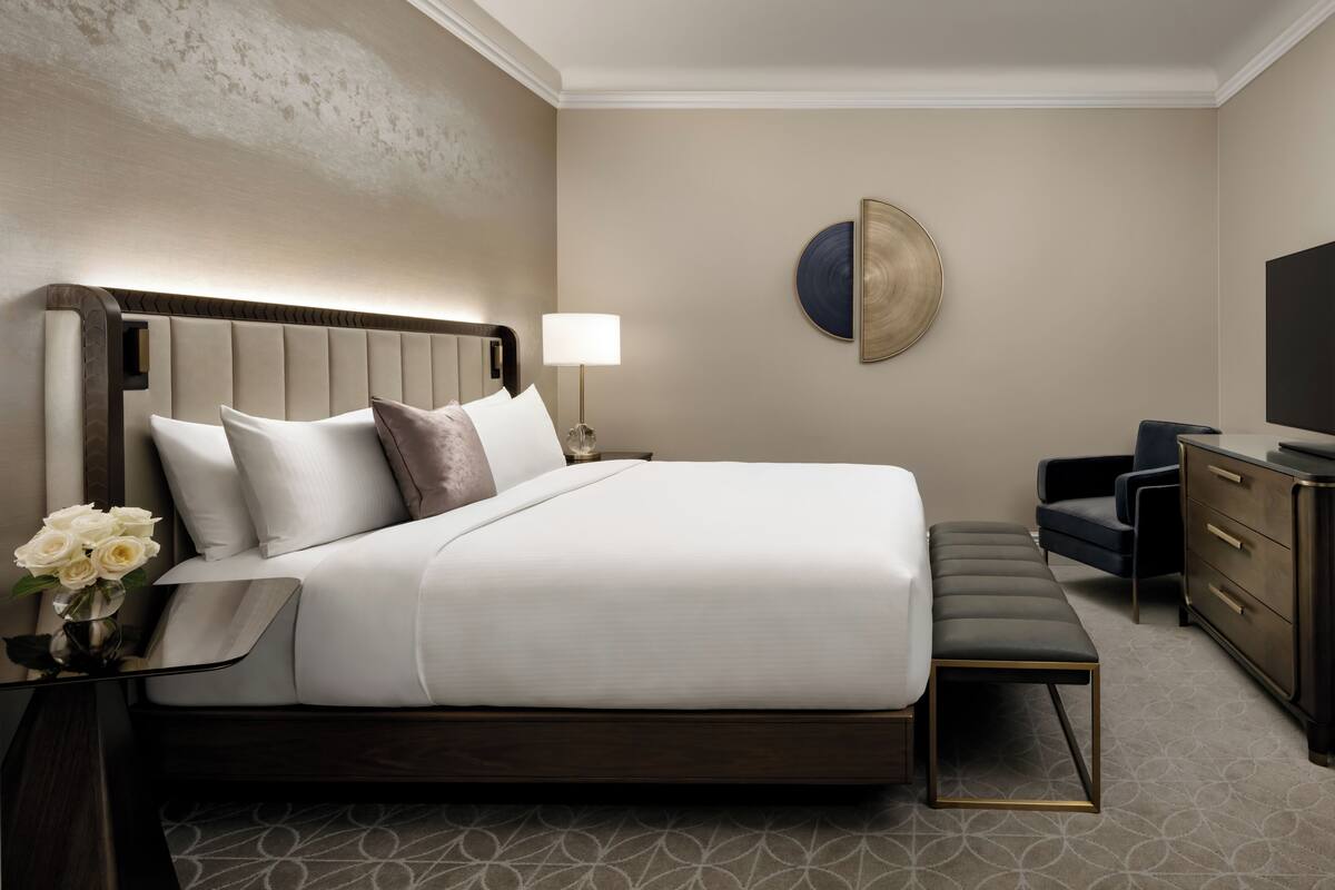 A modern hotel room with a large bed, a bedside table with a lamp, a desk with a chair, and a TV.