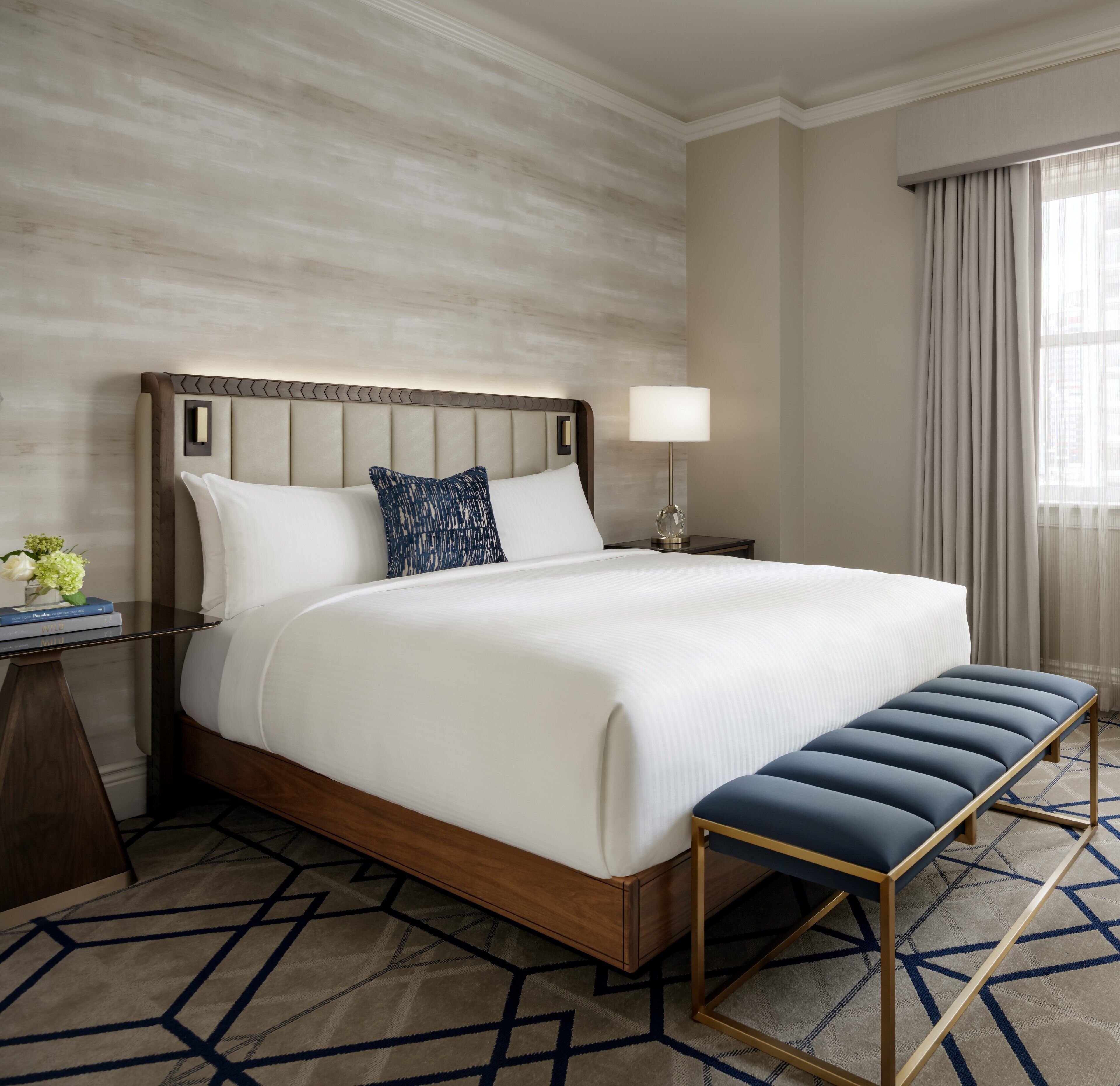 Habitación de hotel con una cama grande, una mesita de noche con lámpara, un banco y un tapete estampado.