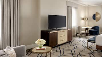 A hotel room with a sofa, a TV, a small table with a vase of flowers, a desk with a lamp, and a chair.