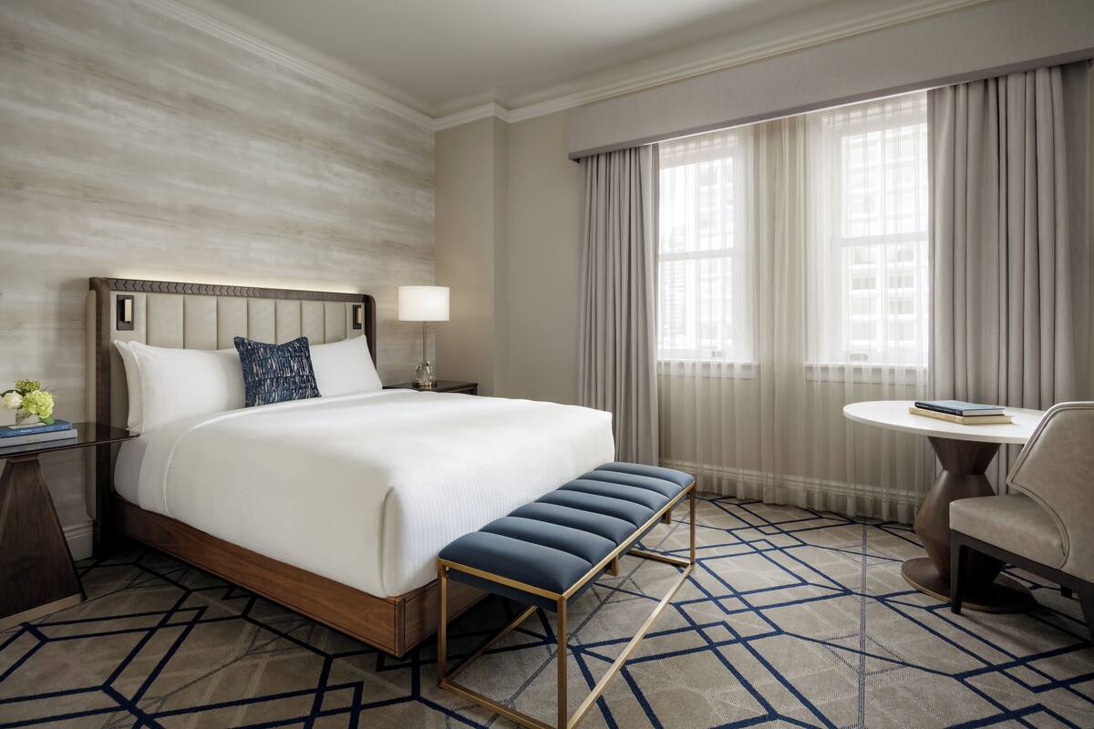 A hotel room with a large bed, a bench, a round table, and a view of the outside through large windows.