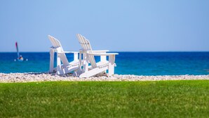 Two white Adirondack chairs on a pebbled beach with a clear blue sky and ocean in the background.