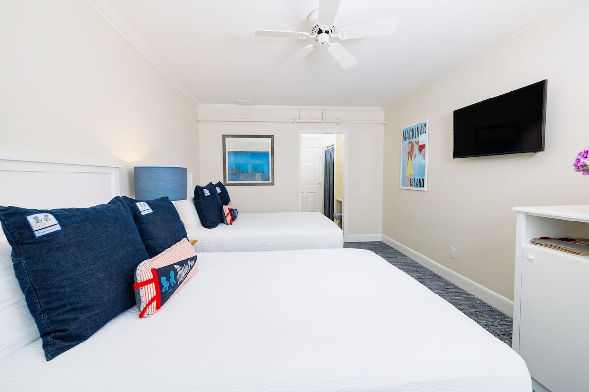 A hotel room with two beds, a ceiling fan, a TV, and a painting on the wall.